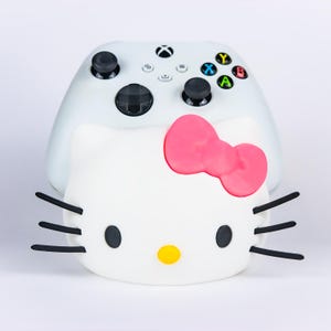 May include: A white video game controller stand shaped like the face of a cartoon cat. The stand features a bright pink bow, black whiskers, two black eyes, and a yellow nose. The controller is white with black joysticks and colored buttons.