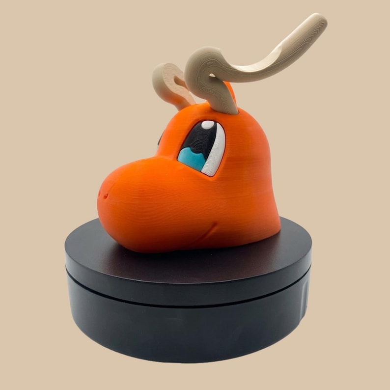 Puede incluir: Una figura de cabeza de criatura naranja impresa en 3D con grandes ojos blancos y azules y dos cuernos beige curvados en la parte superior. La cabeza est&aacute; montada sobre una base negra circular. La superficie muestra capas de impresi&oacute;n 3D visibles.