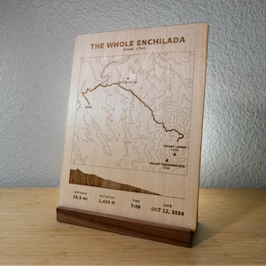 May include: A light-colored wooden plaque featuring a laser-engraved map of The Whole Enchilada trail in Moab, Utah. The plaque displays topographic lines, trail details, and stats including 33.5 mi distance, 1,439 ft elevation, and Oct 12, 2024.