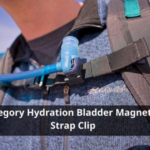 Op de afbeelding: Een blauwe hydratatieblaasslang en clip bevestigd aan een rugzakriem. De clip is zwart en de slang is verbonden met een blauw mondstuk. De tekst "Gregory Hydration Bladder Magnetic Strap Clip" wordt weergegeven.