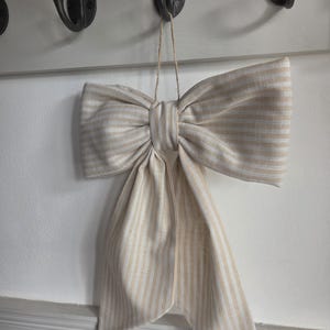 May include: A decorative bow made of fabric with a light beige and white vertical striped pattern. The bow features two large loops and two long, hanging tails, suspended by a thin piece of twine attached to the centre knot of the bow.