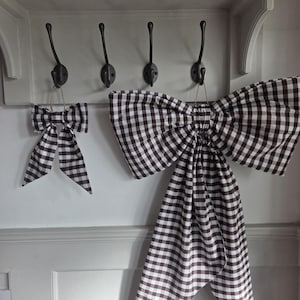 May include: Two decorative bows made of fabric with a brown and white gingham check pattern. Each bow features a central knot and two long, pointed tails hanging downwards. The bows are suspended by thin twine loops from black metal wall hooks.
