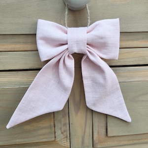 May include: A large, pale pink fabric bow with long, flowing tails. The bow is made of a textured material and is tied in the centre. It is hanging from a white, round knob with a natural fibre string, against a wooden background.