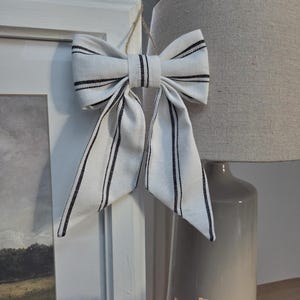 May include: A white fabric bow with black vertical stripes, suspended by twine. The bow is positioned in front of a white picture frame and a grey lamp, creating a decorative accent.