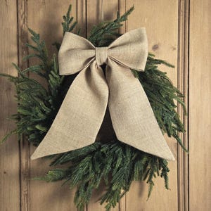 May include: A festive Christmas wreath featuring a large bow made from beige burlap. The wreath is composed of green fir branches and is mounted on a wooden door. The bow is centrally positioned.