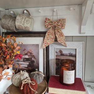 May include: Two beige mugs with the text Hello Autumn, a floral fabric bow, three crocheted pumpkins, a brown glass candle labelled Caramel Waffle Latte, and a book titled Witchcraft & Wizardry arranged on a shelf with framed art prints.