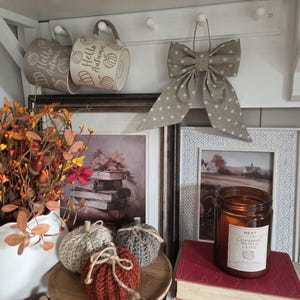 May include: Two beige mugs with Hello Autumn text and pumpkin graphics, a beige polka dot fabric bow, three small crocheted pumpkins, a brown glass candle labelled Caramel Waffle Latte, and two framed prints featuring books and a landscape.