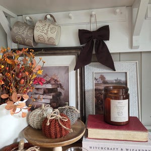 May include: A collection of autumn decor featuring two beige mugs with Hello Autumn text, a dark brown fabric bow, three crocheted pumpkins in rust and grey, a candle labelled Caramel Waffle Latte, and books titled Witchcraft & Wizardry.
