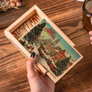May include: A wooden matchbox with a Christmas-themed illustration and the text "Merry Christmas" and "Family Of Charlotte." The box is filled with matches, and a lit match is held near a candle in a glass jar.