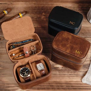 Puede incluir: Estuches de viaje de cuero marrón, uno abierto con relojes y joyas, y dos cerrados. Un estuche cerrado es negro con "NC" en dorado, el otro es marrón con "MC" en dorado. Hay puros en el fondo.