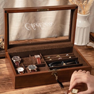 May include: A wooden watch and jewelry box with a glass top. The interior is lined with brown fabric and features compartments for watches, rings, and glasses. Several watches are displayed inside the box. The box is locked with a key.