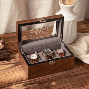 May include: A wooden watch box with a glass top, holding three watches with leather straps. The box is open, revealing a gray interior. The glass top has the initials "C.M.2" etched on it. The box is on a wooden surface.