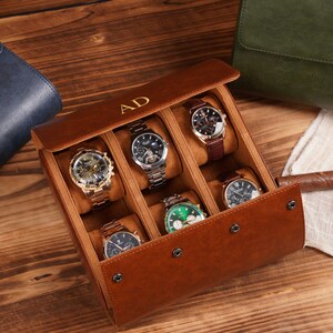 May include: A brown leather watch case with six compartments, each holding a different wristwatch. The case has a hinged lid with the initials "AD" in gold. The watches feature various band and face colors, including gold, silver, black, and brown.