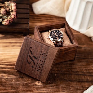 Puede incluir: Una caja de reloj de madera con el nombre "KELLAN" grabado en la tapa. En el interior, un reloj con correa de cuero marrón y esfera negra descansa sobre una tela beige. La caja del reloj está abierta, revelando el reloj. El fondo es una superficie de madera.