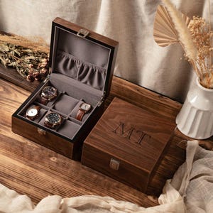 Puede incluir: Una caja de madera de color marr&oacute;n oscuro para relojes con tapa abatible y cierre de metal plateado. El interior est&aacute; forrado con tela gris y tiene seis compartimentos, tres de los cuales contienen relojes. La tapa est&aacute; grabada con MT.