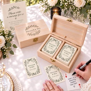 May include: A light-colored wooden box with a hinged lid containing two stacks of cards, each printed with a floral wreath, the names William & Mia, and the date 02.03.2026. A matching sign reads Please write a message for us so we can remember this special day always.