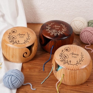 Peut inclure: Trois bols à laine ronds en bois avec des découpes en spirale pour faire passer le fil. Deux bols sont en bois clair avec une gravure de couronne florale, le prénom Anna et la date 05.05. Un bol est en bois foncé avec une rose gravée, Anna et 05.05.
