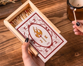 Puede incluir: Una caja de cerillas de madera con una tarjeta decorativa con un diseño de llama, iniciales y el año 2025. La caja de cerillas está abierta, revelando las cerillas en su interior. Una cerilla encendida se sostiene cerca de una vela en un tarro de cristal marrón.