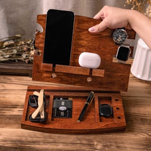 May include: A wooden desk organizer with a dark stain, holding a smartphone, wireless earbuds, a watch, and a smartwatch. The lower section contains sunglasses, a credit card, a pen, and a small black item.