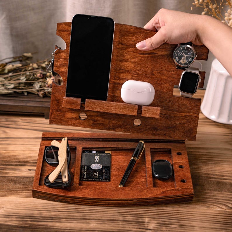 May include: A wooden desk organizer with a dark stain, holding a smartphone, wireless earbuds, a watch, and a smartwatch. The lower section contains sunglasses, a credit card, a pen, and a small black item.