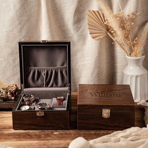 Puede incluir: Una caja de madera de color marr&oacute;n oscuro para relojes con un cierre de metal plateado. La caja abierta tiene un interior de terciopelo gris con tres almohadillas y un bolsillo. La tapa cerrada tiene grabado el nombre William en letra serif.