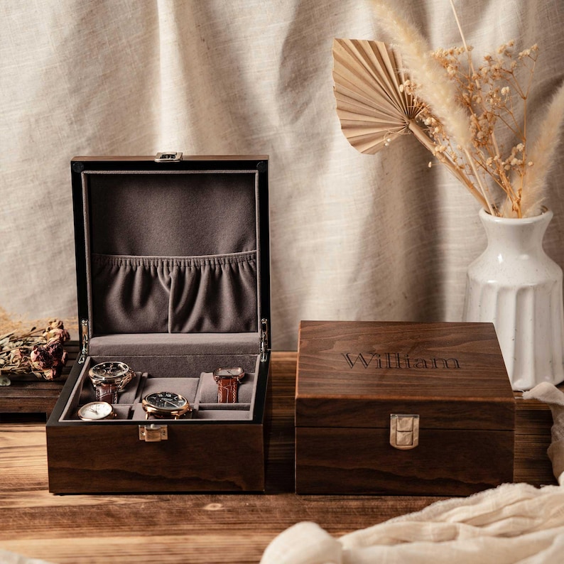 Puede incluir: Una caja de madera de color marr&oacute;n oscuro para relojes con un cierre de metal plateado. La caja abierta tiene un interior de terciopelo gris con tres almohadillas y un bolsillo. La tapa cerrada tiene grabado el nombre William en letra serif.