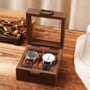 May include: A dark wood watch box with a glass lid featuring the engraved text: Lauren, Love, My Always & Forever, Every Second With You Counts, Rick. The box contains two wristwatches, one with a dark brown leather strap and one with a black strap.