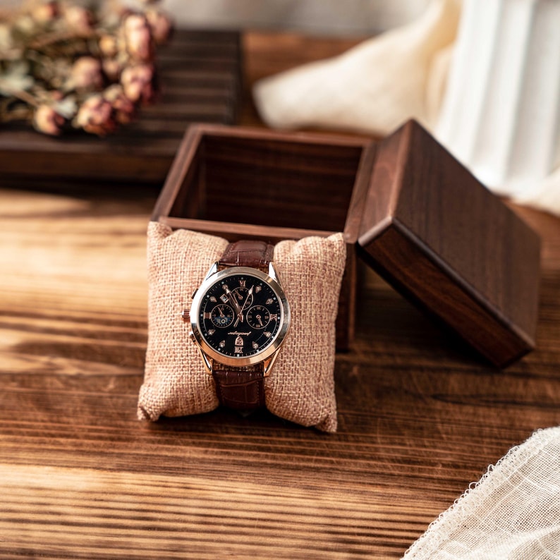 Puede incluir: Un reloj de pulsera con esfera oscura, bisel en tono oro rosa y correa de cuero marr&oacute;n, presentado sobre un coj&iacute;n de arpillera beige dentro de una caja de madera oscura abierta. El reloj cuenta con varias subesferas e indicadores de hora.