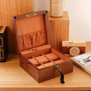 Puede incluir: Una caja de madera de color marr&oacute;n oscuro para relojes con tapa abatible y cerradura de llave de lat&oacute;n. El interior est&aacute; forrado con tela naranja suave, con un bolsillo en la tapa y seis compartimentos con almohadillas. La caja mide 17 cm por 17 cm.