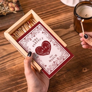 May include: A wooden matchbox with a decorative red and white label that reads "THE PERFECT MATCH" with a heart design and the initials "A + E" and the year "2025". The matchbox is open, revealing the matches inside. A lit match is held near a candle.