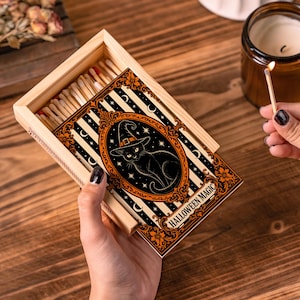 May include: A rectangular wooden matchbox containing wooden matches with red and white tips. The sliding cover features a black cat wearing a witch hat inside an ornate orange frame, set against black and white stripes with the text HALLOWEEN MAGIC.