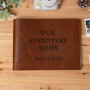May include: A brown leather adventure book with the text "OUR ADVENTURE BOOK" and the names "Katie & Andy" printed in black. A white instant camera and other decorative items are in the background.