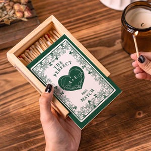 May include: A wooden matchbox with a decorative green and white card that reads "The Perfect Match 2025" with a heart design. The matchbox is open, revealing the matches inside. A lit match is held near a candle.