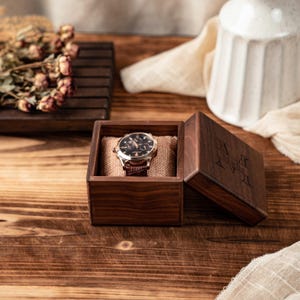 Puede incluir: Una caja de madera cuadrada de color marr&oacute;n oscuro con una tapa abatible que presenta el texto grabado Madie. Dentro de la caja, un reloj de pulsera anal&oacute;gico redondo con esfera oscura y correa de cuero marr&oacute;n descansa sobre un coj&iacute;n de arpillera.