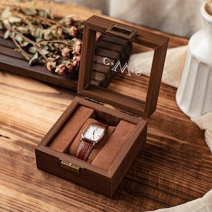 May include: Open wooden watch box with a glass lid, displaying a brown leather-banded watch. The box has a brown velvet interior and brass-colored hardware. The initials "C.M.2" are etched on the glass. The box is on a wooden surface.