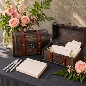 Pode incluir: Dois baús de tesouro de madeira escura com tiras de couro castanho e fechos de metal. Um baú está fechado, com o texto dourado "Lucas & Charlotte" e "16th May 2024". O outro está aberto, contendo cartões de "advice and wishes".