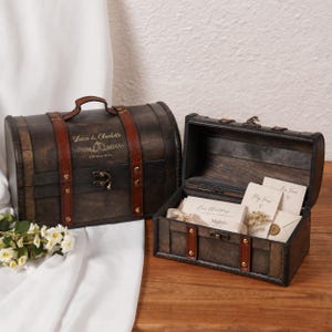 May include: Two dark brown wooden treasure chests with arched lids and brown leather-like straps featuring gold metal studs and latches. The closed chest is engraved with "Lucas & Charlotte" and "12th May 2024". The open chest contains wedding vow cards.