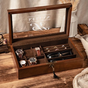 Puede incluir: Una caja de joyería de madera con tapa de cristal, que muestra relojes, anillos y gafas. El interior está forrado con tela marrón. La tapa de cristal tiene el texto "KEITIV" y un símbolo decorativo.