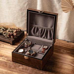 Puede incluir: Una caja para relojes de madera oscura con tapa abatible y cierre met&aacute;lico. El interior est&aacute; forrado con tela gris y cuenta con seis compartimentos individuales, tres de los cuales contienen relojes de pulsera con correas de cuero.