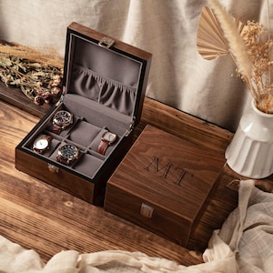 Może przedstawiać: Drewniane pudełko na zegarki w ciemnym kolorze, otwarte, aby odsłonić pięć zegarków z paskami ze skóry. Pudełko jest wyłożone szarą tkaniną i ma srebrne zapięcie. Zamknięte pudełko obok ma wygrawerowane inicjały "MT" na pokrywie.