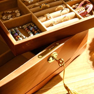 May include: A wooden jewelry box with a key lock and a drawer open, revealing compartments filled with necklaces, bracelets, and rings. The box is made of light brown wood and has a polished finish.