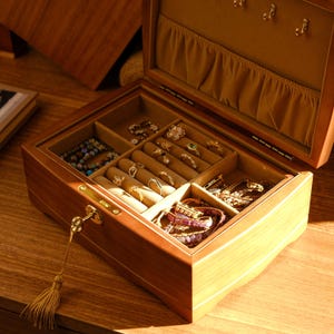 May include: A wooden jewelry box with a key lock and multiple compartments for storing rings, bracelets, and necklaces. The box is open and the interior is lined with brown velvet.