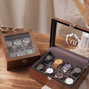 May include: Two wooden watch display cases, one open and one closed, each holding multiple wristwatches. The open case displays watches with various band colors and face designs. The closed case has the initials "CWS" on the glass. The open case has the initials "VB".