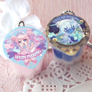 May include: Two small, colorful pudding cups with decorative lids. One is pink with a cartoon girl and the text "WHIM PUDDING". The other is blue with a celestial character and the text "STELLAR PUDDING". Both have keychains.