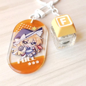 May include: An acrylic keychain featuring an oval charm with an orange gradient background and a cartoon character wearing a witch hat. The text FULL and three stars are printed on the charm. A separate square keycap charm with the letter F is attached.