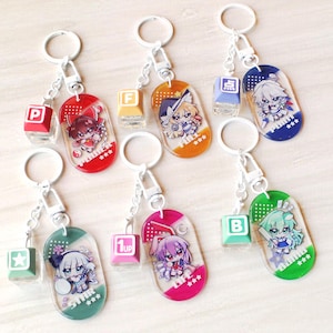 May include: Six acrylic keychains featuring anime-style characters on oval charms with matching square keycap charms. The charms display text including P, F, 1UP, B, and Japanese characters. Each keychain has a silver metal ring and a lobster clasp.