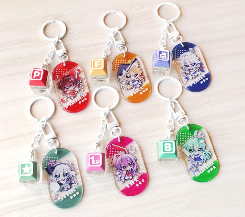 May include: Six acrylic keychains featuring anime-style characters on oval charms with matching square keycap charms. The charms display text including P, F, 1UP, B, and Japanese characters. Each keychain has a silver metal ring and a lobster clasp.