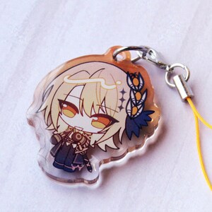May include: A clear acrylic charm featuring a chibi-style anime character with blonde hair, orange eyes, and a dark blue outfit. The character has a white halo and a blue floral accessory. A silver metal lobster clasp attaches to a yellow cord.