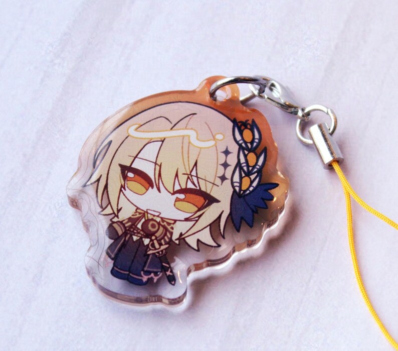May include: A clear acrylic charm featuring a chibi-style anime character with blonde hair, orange eyes, and a dark blue outfit. The character has a white halo and a blue floral accessory. A silver metal lobster clasp attaches to a yellow cord.