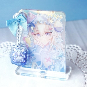 May include: A rectangular acrylic standee featuring a light-haired anime character with purple eyes, decorated with various stickers including a trophy and bowl. A light blue ribbon and a small, dangling acrylic charm are attached to the side.